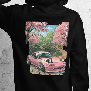 May include: Black hoodie with a graphic of a pink car in a landscape with cherry blossom trees and a bridge. The design includes the text "Pangaea" in a vertical font on the right side of the graphic.