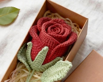 Può includere: Una rosa all'uncinetto rosso scuro con foglie e stelo verde chiaro, inserita in una scatola regalo rettangolare di cartone marrone riempita con carta sminuzzata. La rosa presenta una lavorazione a maglia complessa e petali stratificati.