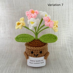 May include: A crocheted potted plant featuring a brown pot with a smiling face, holding a white sign that reads "Votre texte ici Your custom text". The plant has green leaves and several flowers in shades of pink, white, and yellow.