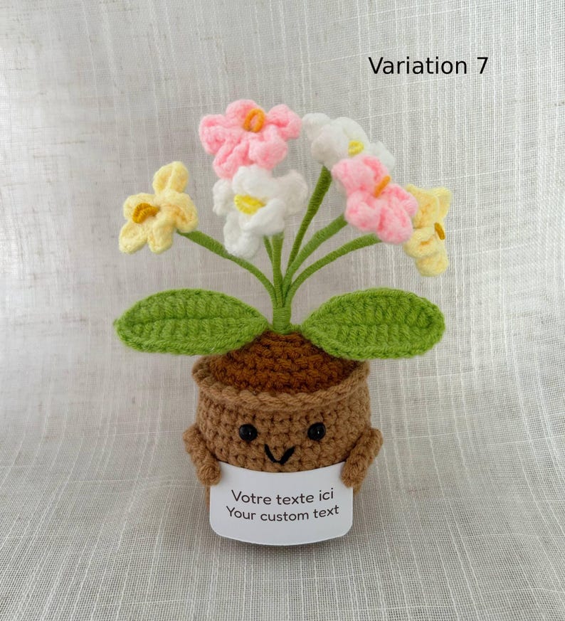 May include: A crocheted potted plant featuring a brown pot with a smiling face, holding a white sign that reads "Votre texte ici Your custom text". The plant has green leaves and several flowers in shades of pink, white, and yellow.