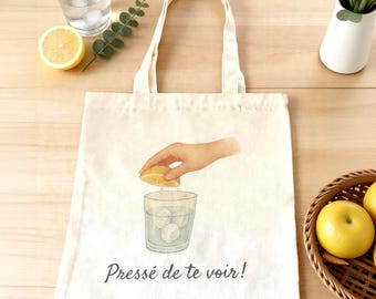 Può includere: Una borsa in tela color crema con un'illustrazione grafica di una mano che spreme uno spicchio di limone in un bicchiere d'acqua con cubetti di ghiaccio. Sotto l'illustrazione, il testo Pressé de te voir è stampato in corsivo grigio scuro.