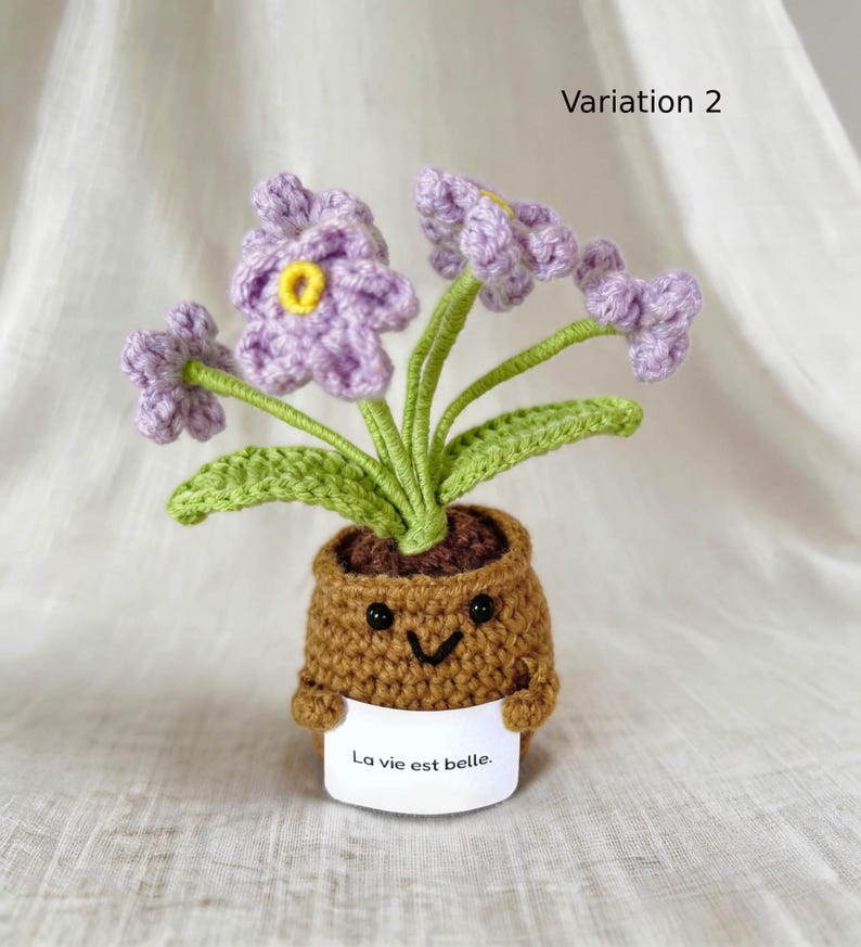 May include: A crocheted potted plant featuring light purple flowers with yellow centers and green leaves. The brown pot has a smiling face and holds a white sign with the text La vie est belle. The item is labeled as Variation 2 in the upper right corner.