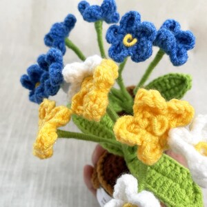May include: A crochet flower arrangement featuring blue, yellow, and white blossoms with five petals each. The flowers have yellow centers and are attached to green stems with textured green leaves. The stems are anchored in a small brown crochet pot.