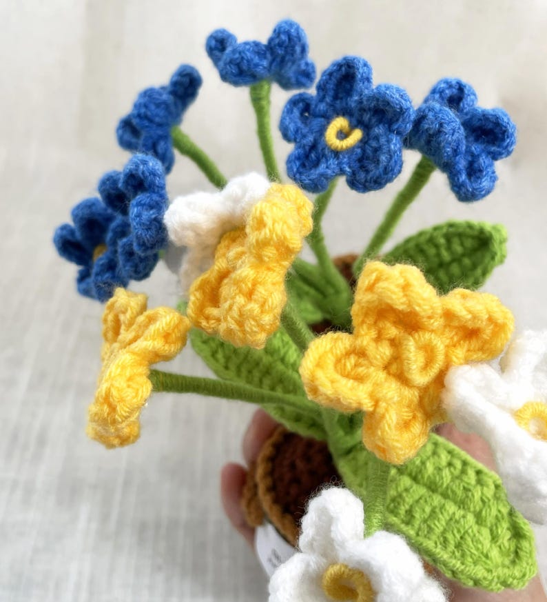 May include: A crochet flower arrangement featuring blue, yellow, and white blossoms with five petals each. The flowers have yellow centers and are attached to green stems with textured green leaves. The stems are anchored in a small brown crochet pot.