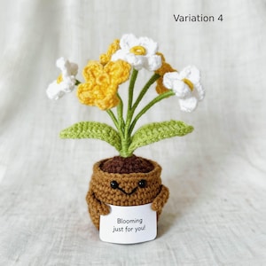 May include: A crocheted potted plant with white and yellow flowers, green stems, and leaves. The brown pot features a stitched smiling face and holds a white sign with the black text Blooming just for you! The image is labeled Variation 4.