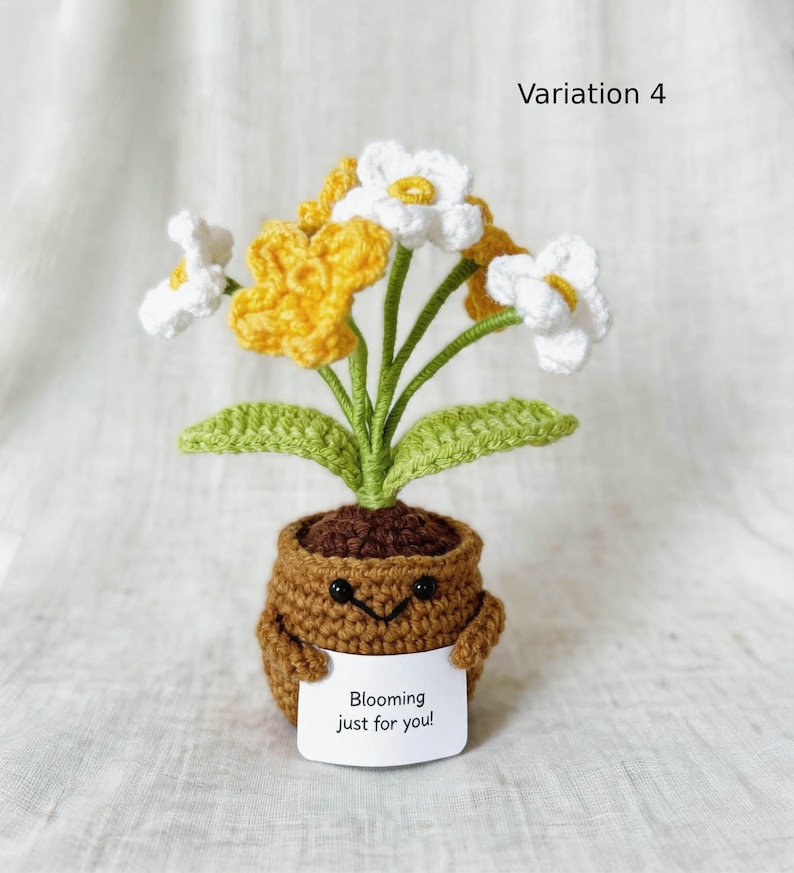 May include: A crocheted potted plant with white and yellow flowers, green stems, and leaves. The brown pot features a stitched smiling face and holds a white sign with the black text Blooming just for you! The image is labeled Variation 4.
