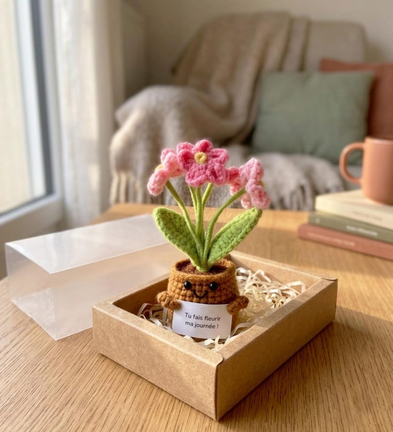 May include: A crocheted potted plant with pink flowers and green leaves in a brown pot with a smiling face. The pot holds a white sign with the text "Tu fais fleurir ma journ&eacute;e !" and is packaged in a cardboard box with a clear plastic lid.