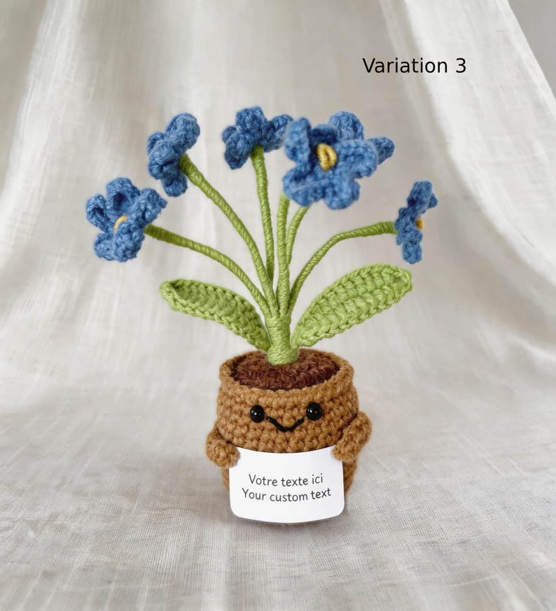 May include: A crocheted potted plant featuring five blue flowers with yellow centers and green stems and leaves. The brown pot has a smiling face and holds a white sign with the text "Votre texte ici Your custom text". Variation 3 is labeled.