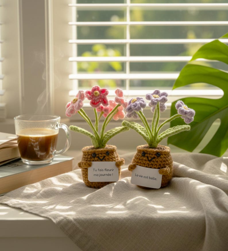 May include: Two crocheted potted plants with smiling faces, one with pink flowers and a sign reading 'Tu fais fleurir ma journ&eacute;e !' and the other with purple flowers and a sign reading 'La vie est belle.', both featuring green stems and brown pots.