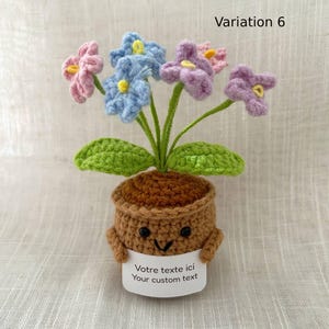 May include: A crocheted potted plant featuring pink, blue, and purple flowers with green stems and leaves. The brown pot has a smiling face with black bead eyes and holds a white sign that reads "Votre texte ici Your custom text".