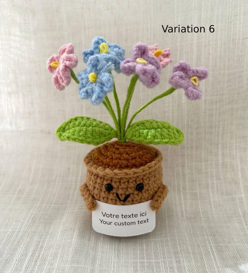 May include: A crocheted potted plant featuring pink, blue, and purple flowers with green stems and leaves. The brown pot has a smiling face with black bead eyes and holds a white sign that reads "Votre texte ici Your custom text".