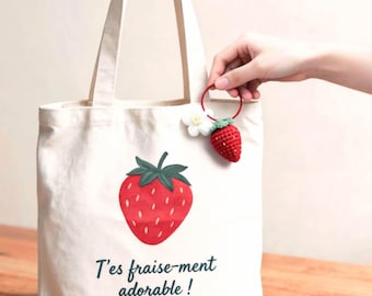 Può includere: Una borsa tote in tela color crema con una grande grafica di una fragola rossa con foglie verdi e il testo "T'es fraise-ment adorable !" stampato in verde scuro. È attaccato un piccolo ciondolo a forma di fragola rossa all'uncinetto.