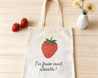 Può includere: Una borsa tote in tela color crema con un'illustrazione centrale di una fragola rossa con foglie verdi. Sotto la fragola, il testo "T'es fraise-ment adorable !" è stampato in un carattere corsivo scuro sulla superficie della borsa.