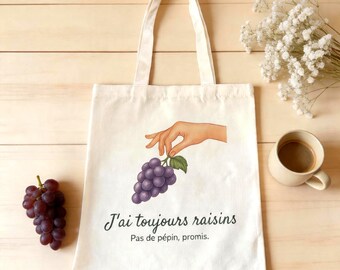 Può includere: Una borsa tote in tela color crema con un'illustrazione grafica di una mano che tiene un grappolo d'uva viola. Il testo "J'ai toujours raisins" e "Pas de pépin, promis." è stampato in nero sotto l'illustrazione dell'uva.