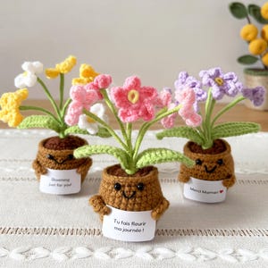 May include: Three crocheted potted plants with smiling brown pots, each holding a white sign. The left sign reads "Blooming just for you!", the center "Tu fais fleurir ma journ&eacute;e!", and the right "Merci Maman" with a red heart symbol.