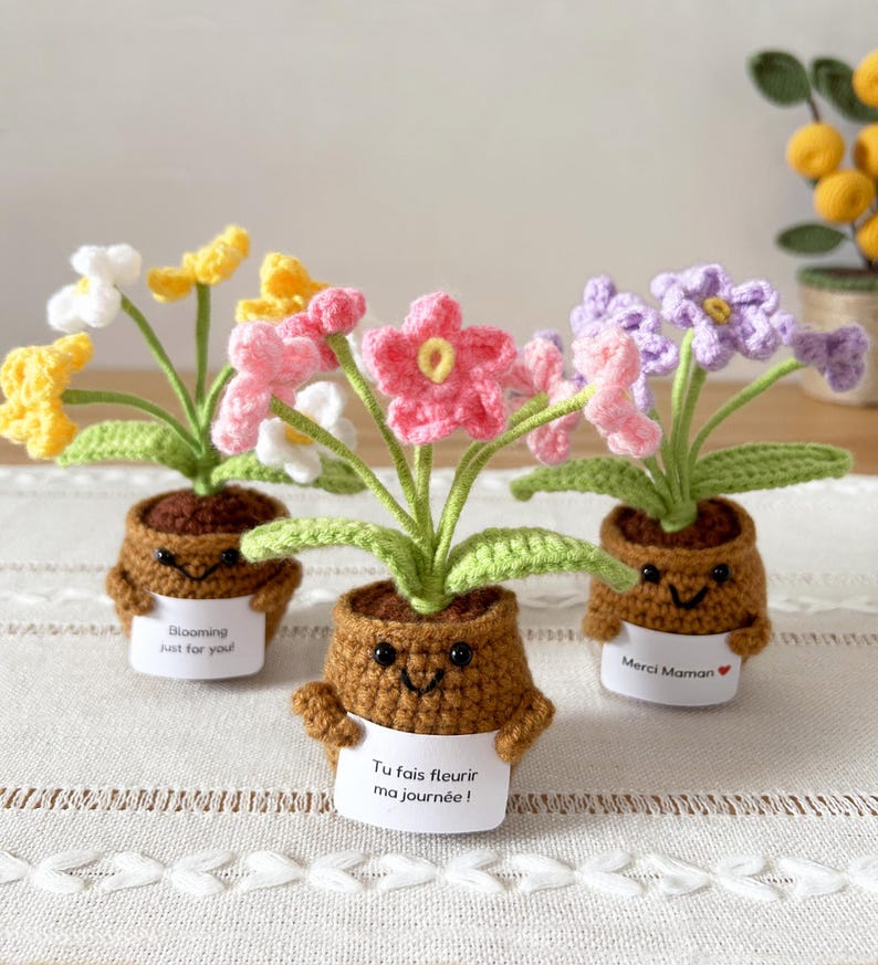 May include: Three crocheted potted plants with smiling brown pots, each holding a white sign. The left sign reads "Blooming just for you!", the center "Tu fais fleurir ma journ&eacute;e!", and the right "Merci Maman" with a red heart symbol.