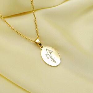 May include: A gold-colored necklace with an oval pendant. The pendant features an engraved floral design. The chain and pendant are a bright, shiny gold. The necklace is displayed on a light yellow fabric.