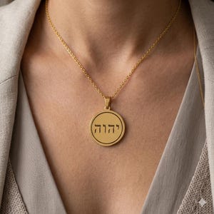 Puede incluir: Un collar con colgante dorado con un dije circular. El dije presenta texto hebreo dentro de un borde circular. El collar se usa sobre un fondo de tonos neutros, resaltando el diseño y el color de la joya.
