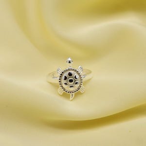 Op de afbeelding: Een zilveren ring met een schildpadmotief. De schildpad heeft een gedetailleerd schild met een textuurpatroon en kleine, ronde accenten. De ring is geplaatst tegen een zachte, gele achtergrond.