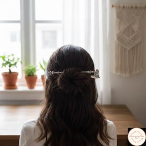 Könnte beinhalten: Ein silberfarbener Haarnadel, geformt wie eine kleine Axt, wird in dunkelbraunem Haar getragen. Die Nadel ist horizontal am Hinterkopf positioniert und sichert einen Dutt. Der Hintergrund umfasst ein Fenster, Pflanzen und einen Makramee-Wandbehang.