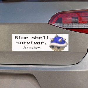 May include: A silver car with a bumper sticker that reads "Blue shell survivor. Ask me how." and features a blue and white cartoon character.