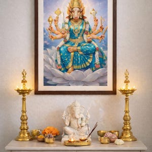 May include: A framed artwork depicting a multi-armed deity in blue and gold attire, seated on a white lotus flower. The image is displayed above a white cabinet with gold lamps, a white statue, and small bowls with flowers.