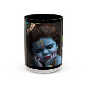 May include: A white ceramic mug with a black interior and a detailed image of a blue-skinned child with a sad expression. The child wears gold jewelry and a jeweled headpiece. The mug has a white base.