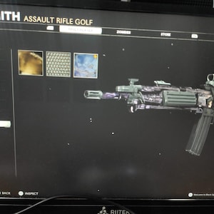 May include: A screen displays the "UNSMITH ASSAULT RIFLE GOLF" weapon customization menu. The image shows a detailed assault rifle with a dark, metallic finish. The screen also shows customization options and the words "ZOMBIES" and "STORE".