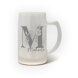 Könnte beinhalten: Ein Bierkrug aus mattiertem Glas mit einem großen grauen Buchstaben "M", der mit floralen Elementen verziert ist. Der Name "Michaela" steht darunter in Schreibschrift. Der Krug hat einen Henkel und einen breiten Boden.