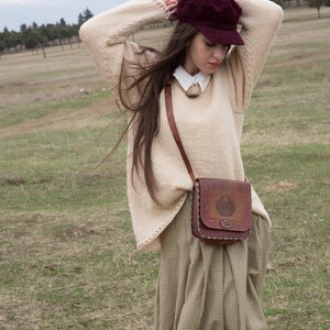 Könnte beinhalten: Eine Person trägt eine bordeauxfarbene Ballonmütze, einen cremefarbenen Pullover und eine braune Leder-Umhängetasche mit geprägtem Design. Der Rock hat ein kariertes Muster in Beige und Braun. Der Hintergrund ist eine Wiese.