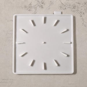 May include: A square white silicone mold for a clock face featuring twelve raised triangular markers arranged in a circular pattern and a small central hole for a clock mechanism. The mold has a smooth finish and a small tab at the top edge.
