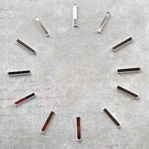 May include: Twelve small, thin, rectangular silver-toned metal bars arranged in a circular clock-like formation. Each individual bar features a smooth, reflective surface and straight edges, positioned evenly to represent the twelve hour markers.