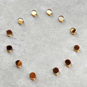 May include: A collection of small, round, gold-colored metal discs arranged in a circular pattern on a light gray surface. The discs have a reflective, polished finish, catching the light and creating highlights.