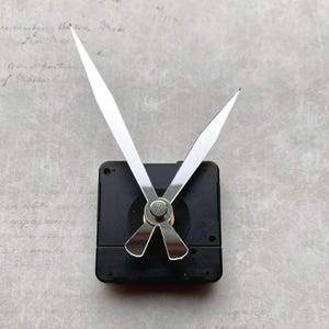 May include: A black clock movement with silver hands. The hands are long and pointed, indicating the time. The clock mechanism is square and has a central spindle. The background is a light gray with a subtle floral and script pattern.