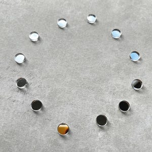 Può includere: Una composizione circolare di piccoli pezzi rotondi di vetro a specchio su una superficie grigia. Le superfici a specchio riflettono l'ambiente, creando effetti di luce e ombra. Un pezzo ha un centro dorato. Ogni pezzo misura circa 1 cm di diametro.