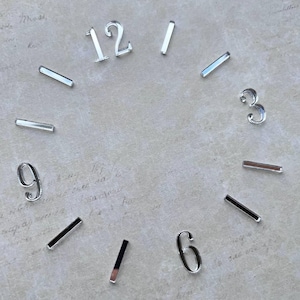 May include: A set of silver-coloured clock numerals and markers arranged in a circular pattern. The set includes the digits 12, 3, 6, and 9, along with eight rectangular bar markers. Each piece features a reflective, metallic finish.