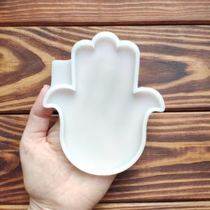 May include: A white, hand-shaped mold, held in a person's hand, against a wooden background. The mold has a smooth surface and a detailed outline of a hand with a thumb and four fingers. The mold is likely used for crafting or decorative purposes.