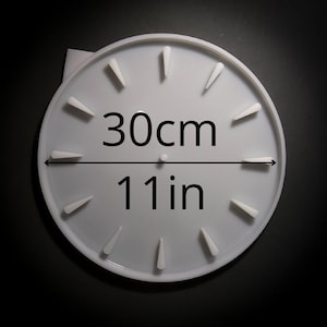 Könnte beinhalten: Ein weißes, kreisförmiges Zifferblatt mit erhabenen, dreieckigen Stundenmarkierungen. Das Zifferblatt hat einen Durchmesser von 30 cm (11 Zoll), wobei die Messung deutlich in schwarzem Text angezeigt wird.