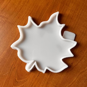 May include: A white, leaf-shaped mold with a jagged edge. The mold is made of a smooth, solid material and is set against a warm, brown wooden surface. The design is simple and clean, with a clear, open center.