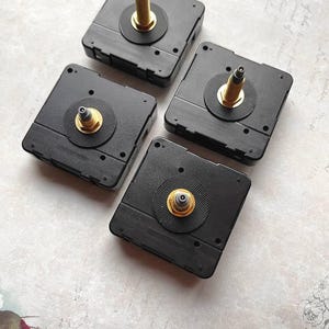May include: Four black clock movements with gold-colored spindles and circular black rubber washers. The square-shaped mechanisms are arranged on a light-colored surface, showcasing their design and components for clock making or repair.