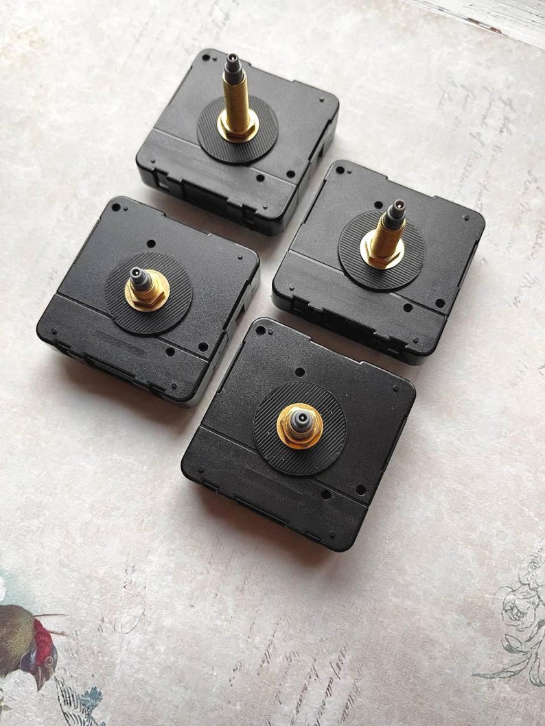 May include: Four black clock movements with gold-colored spindles and circular black rubber washers. The square-shaped mechanisms are arranged on a light-colored surface, showcasing their design and components for clock making or repair.