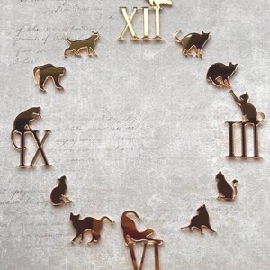 Könnte beinhalten: Ein Set aus goldfarbenen Acryl-Uhrenkomponenten mit zwölf einzelnen Katzensilhouetten in verschiedenen Posen und vier römischen Ziffern XII, III, VI und IX, die kreisförmig angeordnet sind, um ein Zifferblatt zu bilden.