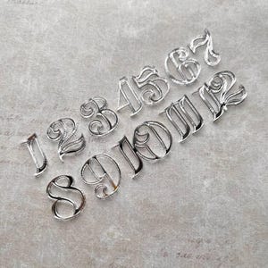 Può includere: Una collezione di numeri in metallo argentato, da uno a dodici, in un carattere decorativo. I numeri sono disposti diagonalmente su una superficie grigio chiaro, ognuno con un design unico e ornato. Adatto per artigianato o decorazioni.
