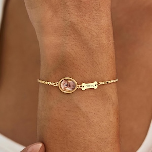 Könnte beinhalten: Ein goldfarbenes Kettenarmband mit einem kleinen ovalen Anhänger mit Tierporträt und einem weiteren Anhänger in Form eines Hundeknochens. Der Knochenanhänger ist mit dem Namen Marley graviert. Das Armband hat ein feines Gliederkettendesign.