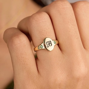 May include: A gold signet ring featuring an oval face engraved with the cursive letter B. The band includes two small star-shaped settings, each containing a single green gemstone, positioned on either side of the central oval plaque.