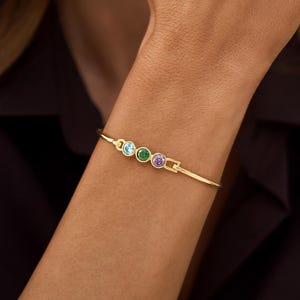 Peut inclure: Un bracelet jonc doré orné de trois pierres précieuses circulaires serties en rangée. Les pierres sont bleu clair, vertes et violettes. Le bracelet présente une fine bande métallique polie et un petit fermoir visible sur le côté.