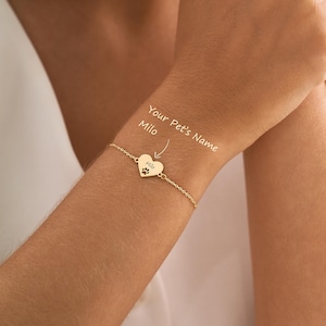 May include: A gold-colored chain bracelet featuring a small heart-shaped charm. The charm is engraved with a paw print and the name Milo. Text on the image indicates Your Pet's Name and points to the charm. The bracelet has a delicate link chain.