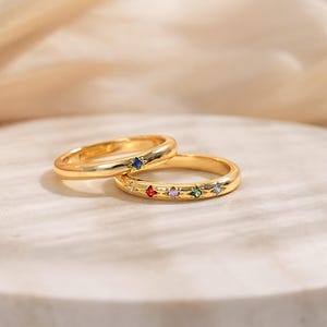 Könnte beinhalten: Zwei goldene Ringe mit sternförmigen Fassungen. Ein Ring hat einen einzelnen blauen Edelstein, während der andere eine Reihe von fünf bunten Edelsteinen in Rot, Lila, Grün und Blau auf einem polierten goldenen Ringband aufweist.