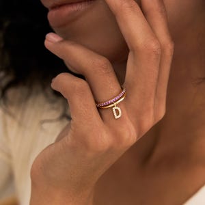 Peut inclure: Une bague dorée composée d'un anneau serti de petites pierres roses et d'une breloque en forme de lettre D, ornée de minuscules pierres transparentes. La bague est portée au doigt, mettant en valeur la finition métallique et les pierres.