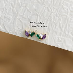 May include: A pair of gold stud earrings featuring a marquise-cut purple stone, a round clear stone, and a rectangular green baguette stone. The text "Your Family or Friend Birthstone" is printed above the earrings with an arrow pointing to them.